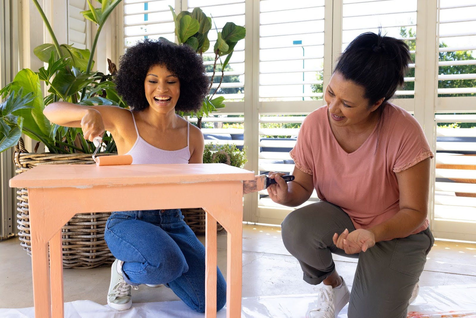 Two female friends smiling while painting thrifted furniture